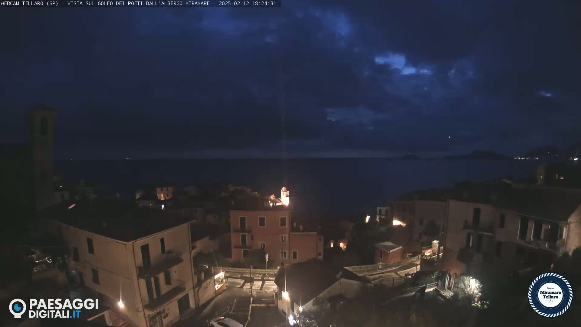 A nighttime view of a coastal town overlooking a dark, calm sea under a partly cloudy sky.