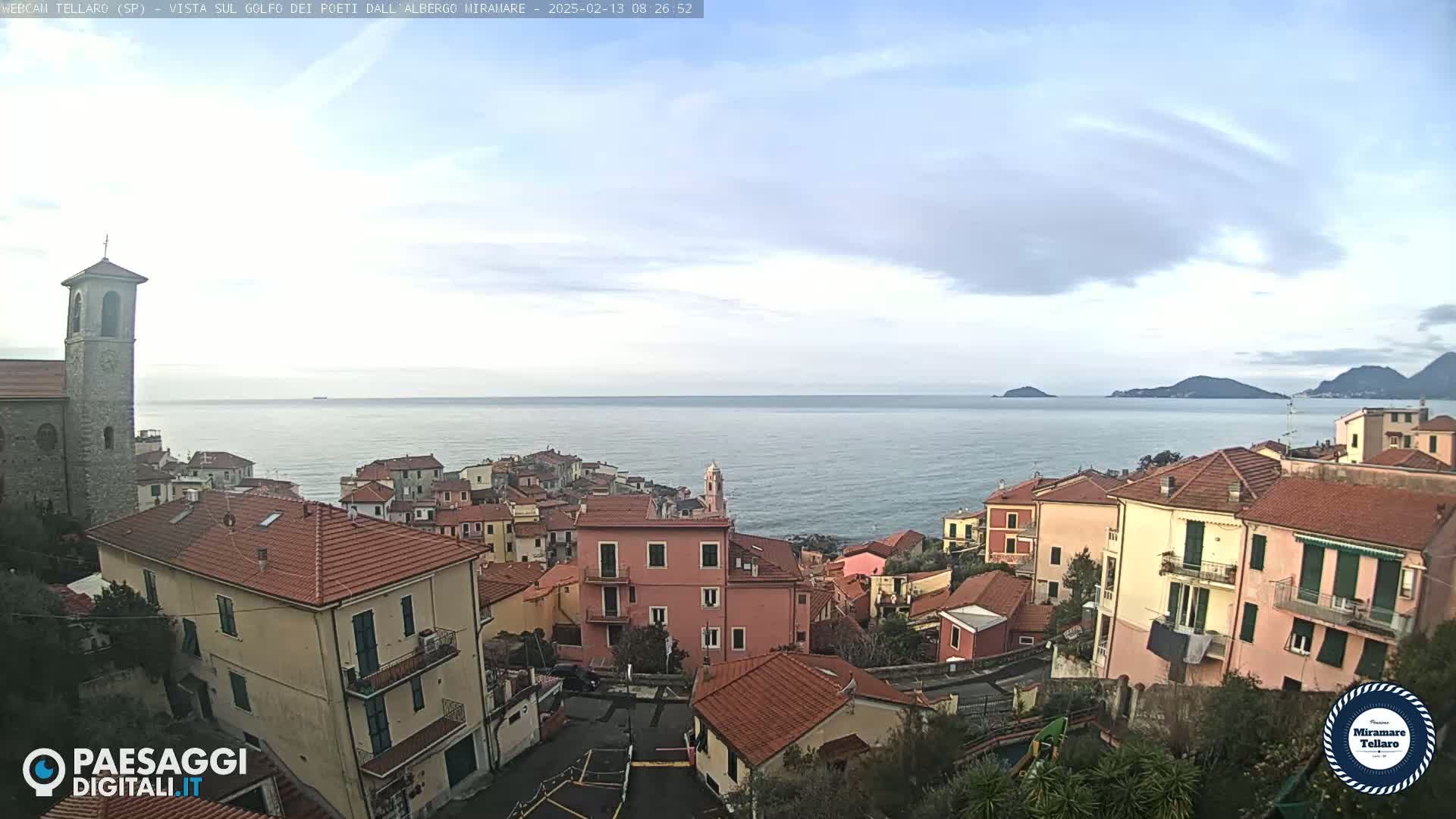 A coastal town with red-tiled roofs overlooks a calm sea under a mostly cloudy sky.