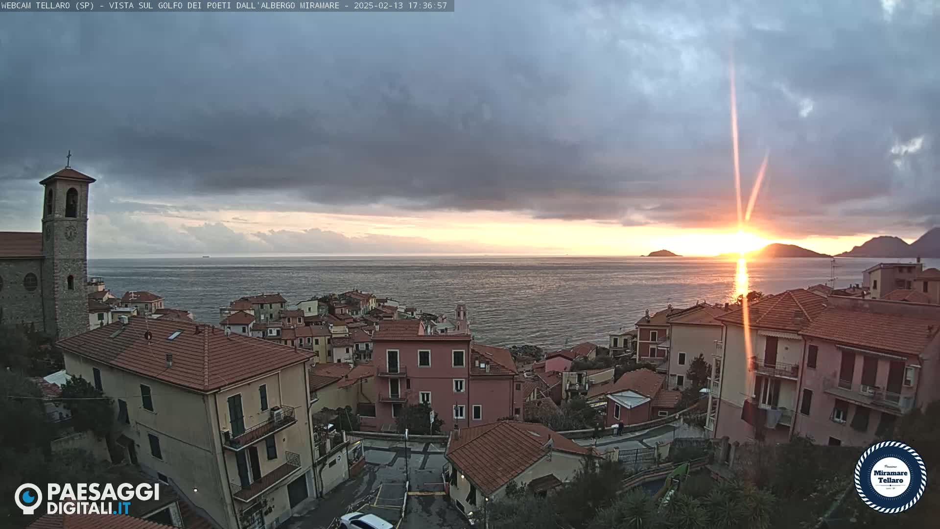 A coastal town with red-tiled roofs sits on a hillside overlooking a calm sea at sunset under a partly cloudy sky.