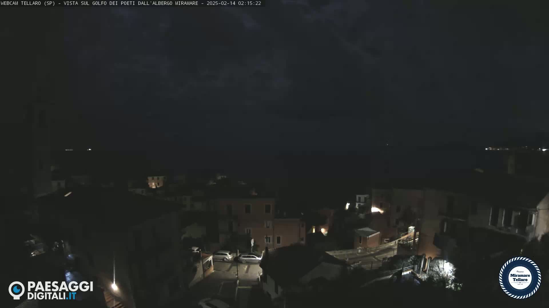 A nighttime view of a coastal town shows dimly lit buildings and streets overlooking a dark body of water under a cloudy sky.
