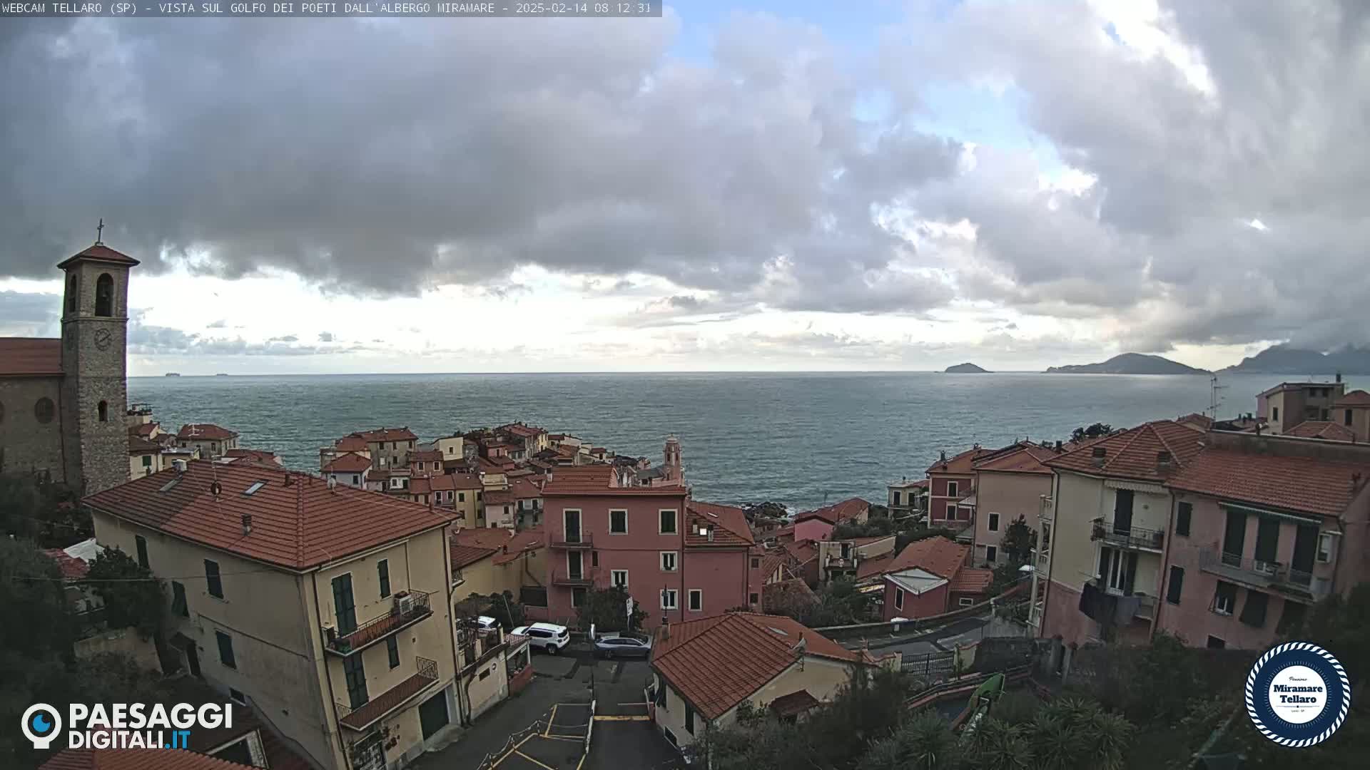 A coastal town with red-tiled roofs and a church steeple overlooks a calm sea under a cloudy sky.