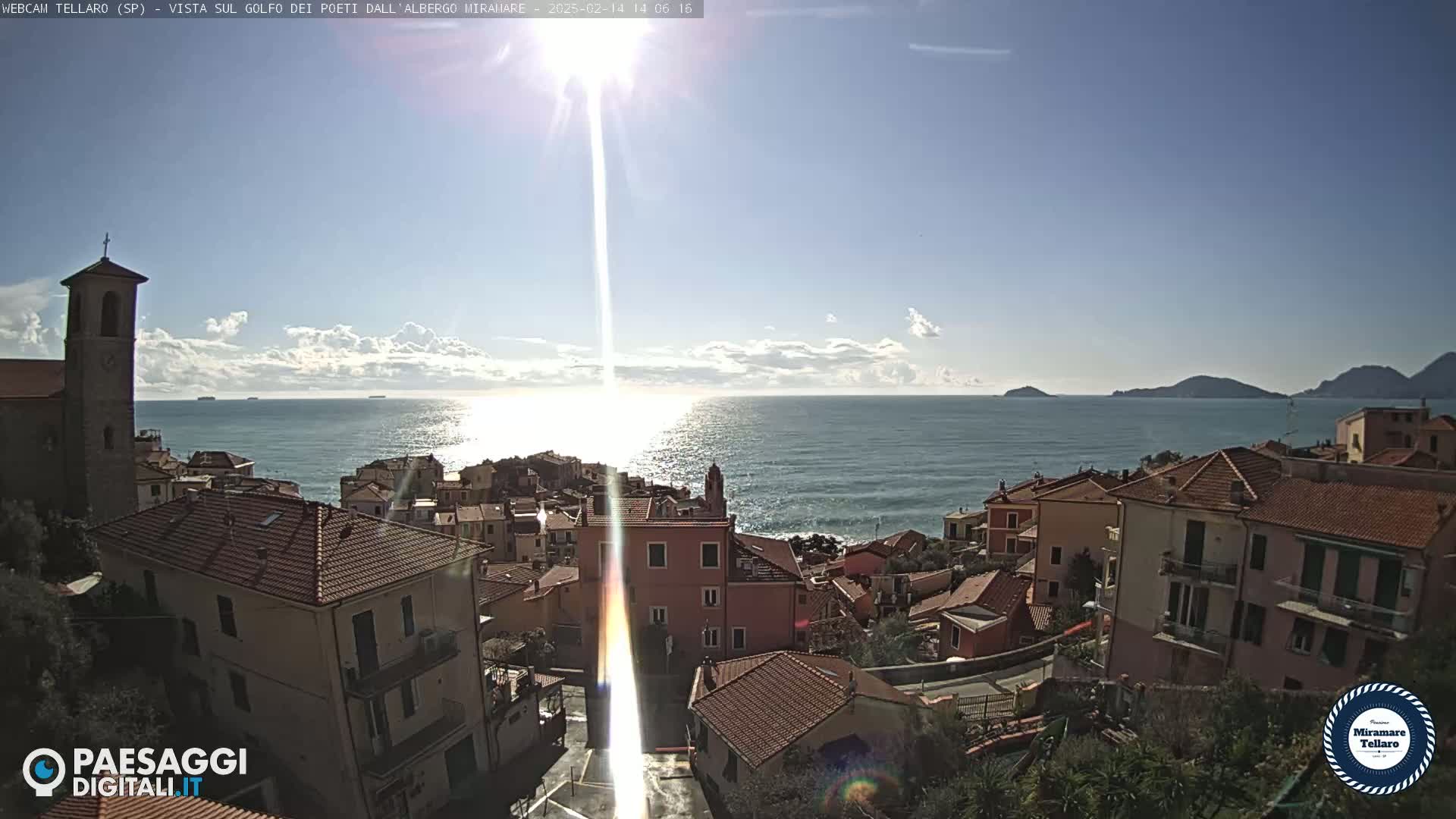 A coastal town with red-tiled roofs and a church steeple overlooks a calm, sunlit sea under a partly cloudy sky.