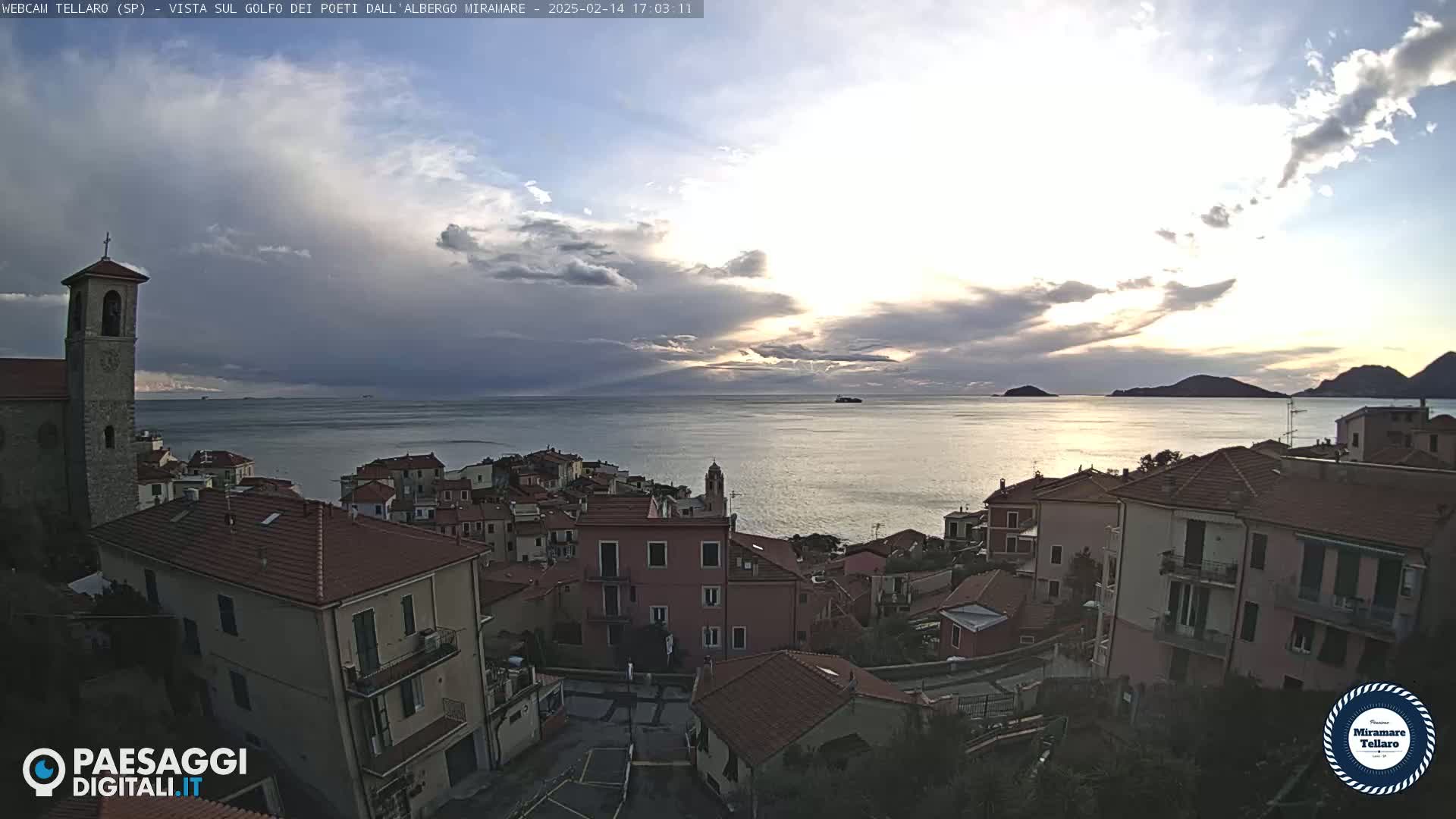 A coastal town with red-roofed buildings overlooks a calm sea under a partly cloudy sky.