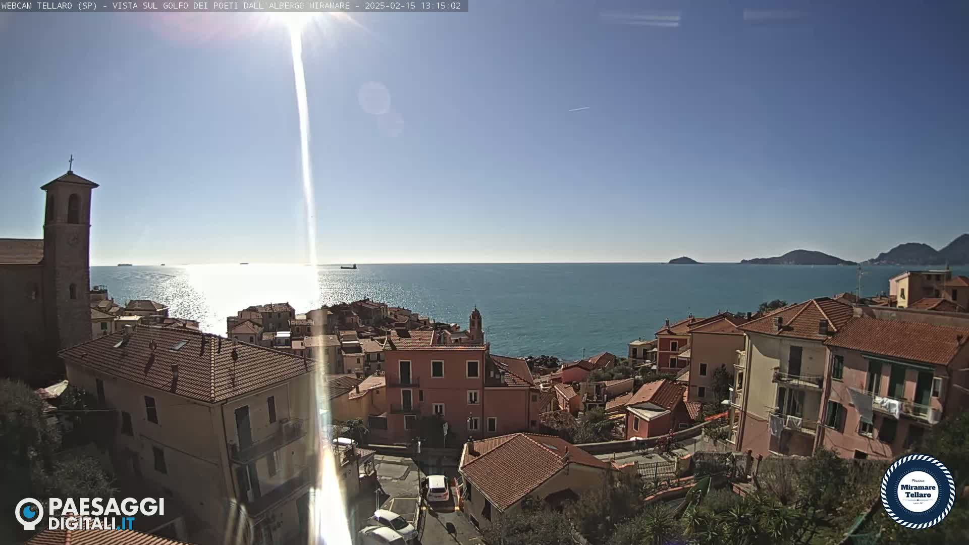 A sunny coastal town with red-tiled roofs overlooks a calm, blue sea under a clear sky.