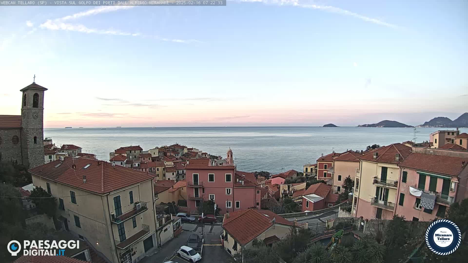 A coastal town with red-tiled roofs overlooks a calm sea under a clear, pale sky at dawn.