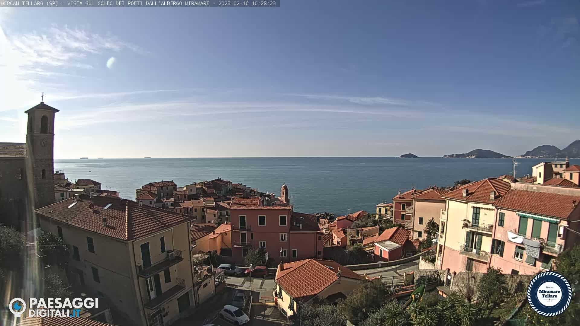 A coastal town with red-tiled rooftops and a church steeple overlooks a calm sea under a mostly clear sky.