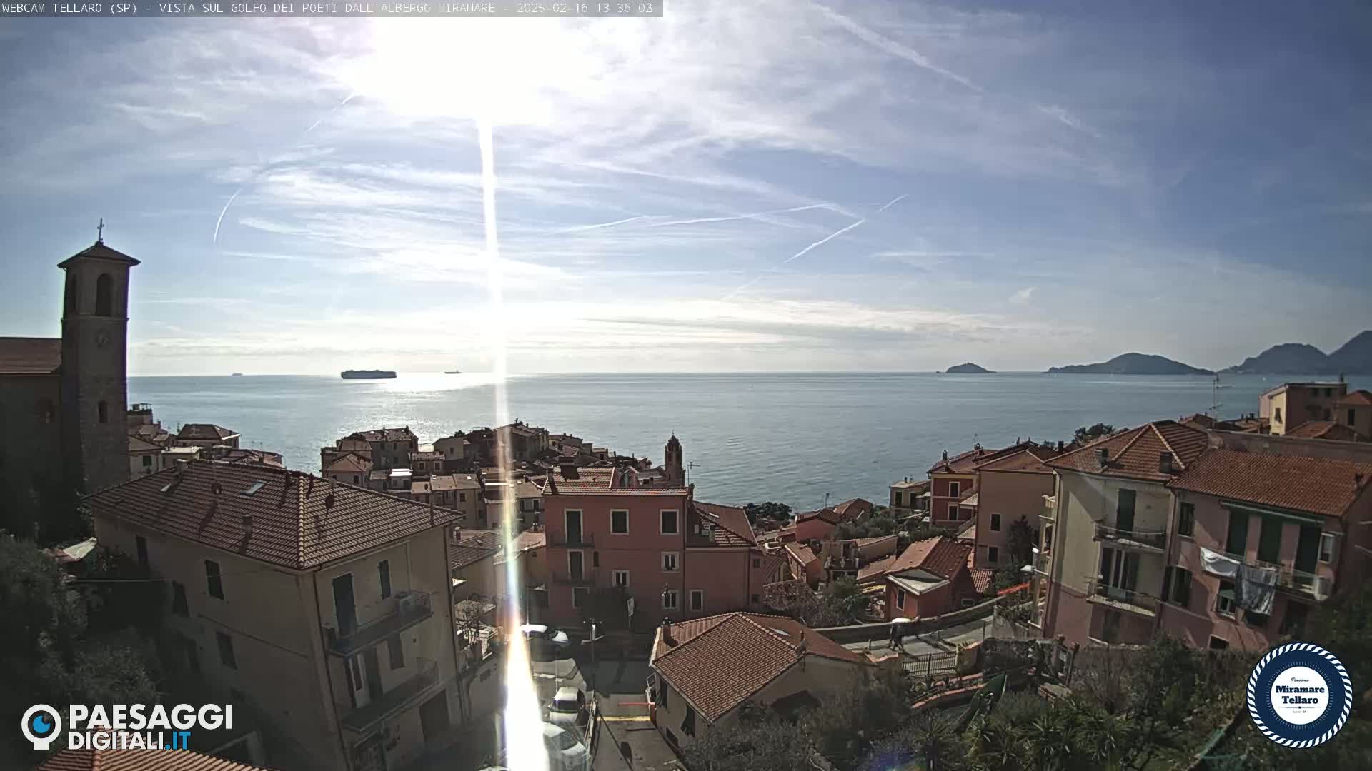 A coastal town with red-tiled rooftops overlooks a calm sea under a mostly sunny sky with a few scattered clouds.