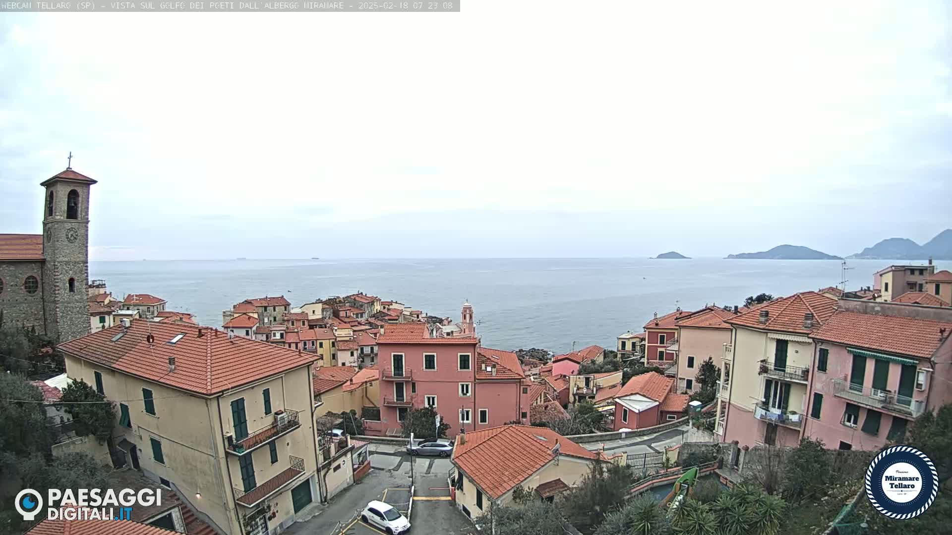 A coastal town with red-tiled roofs, a church steeple, and a calm sea under an overcast sky.