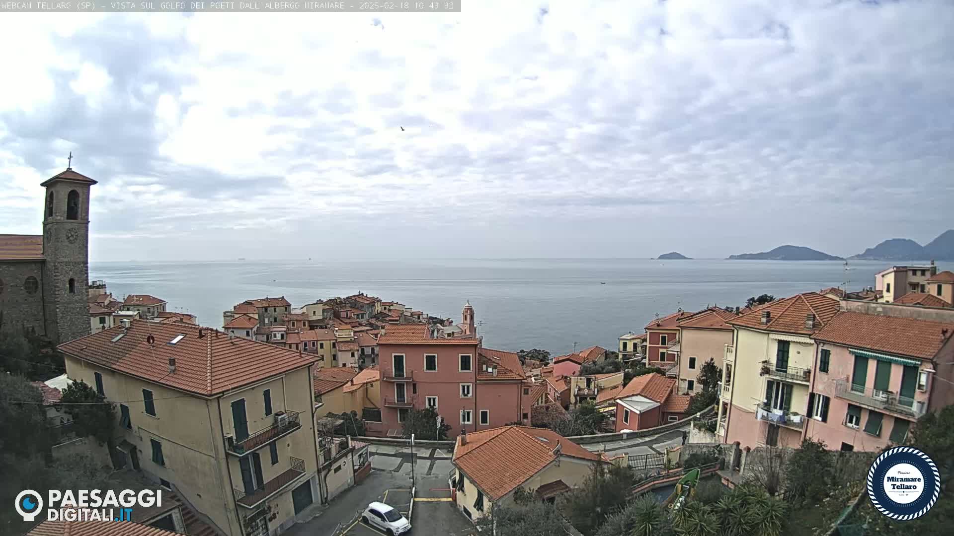 A coastal town with red-tiled rooftops and a stone church overlooks a calm sea under a partly cloudy sky.