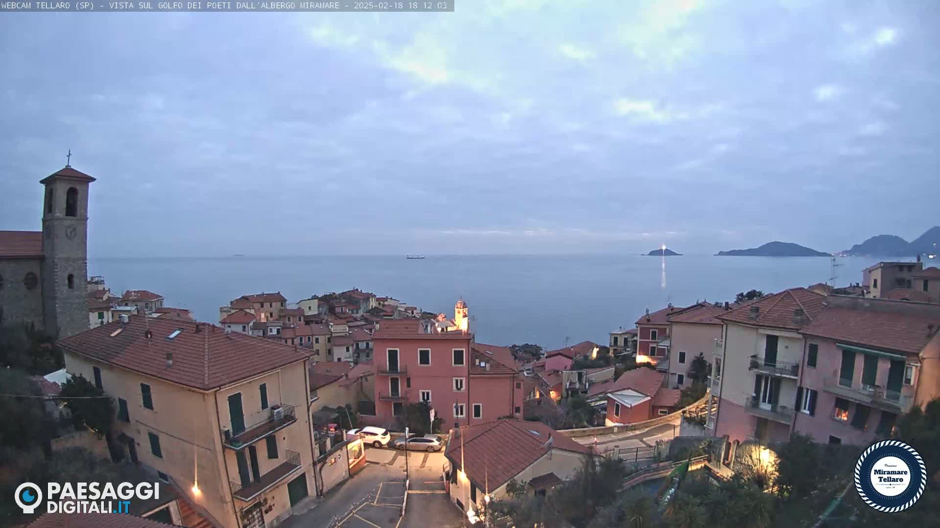 A coastal town with red-roofed buildings overlooks a calm sea under a mostly clear, dusky sky.