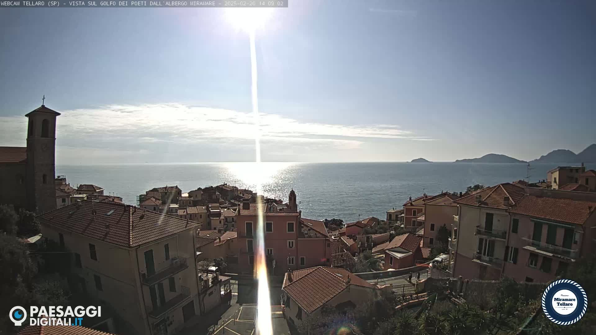 A coastal town with red-tiled roofs overlooks a calm sea under a bright, sunny sky.