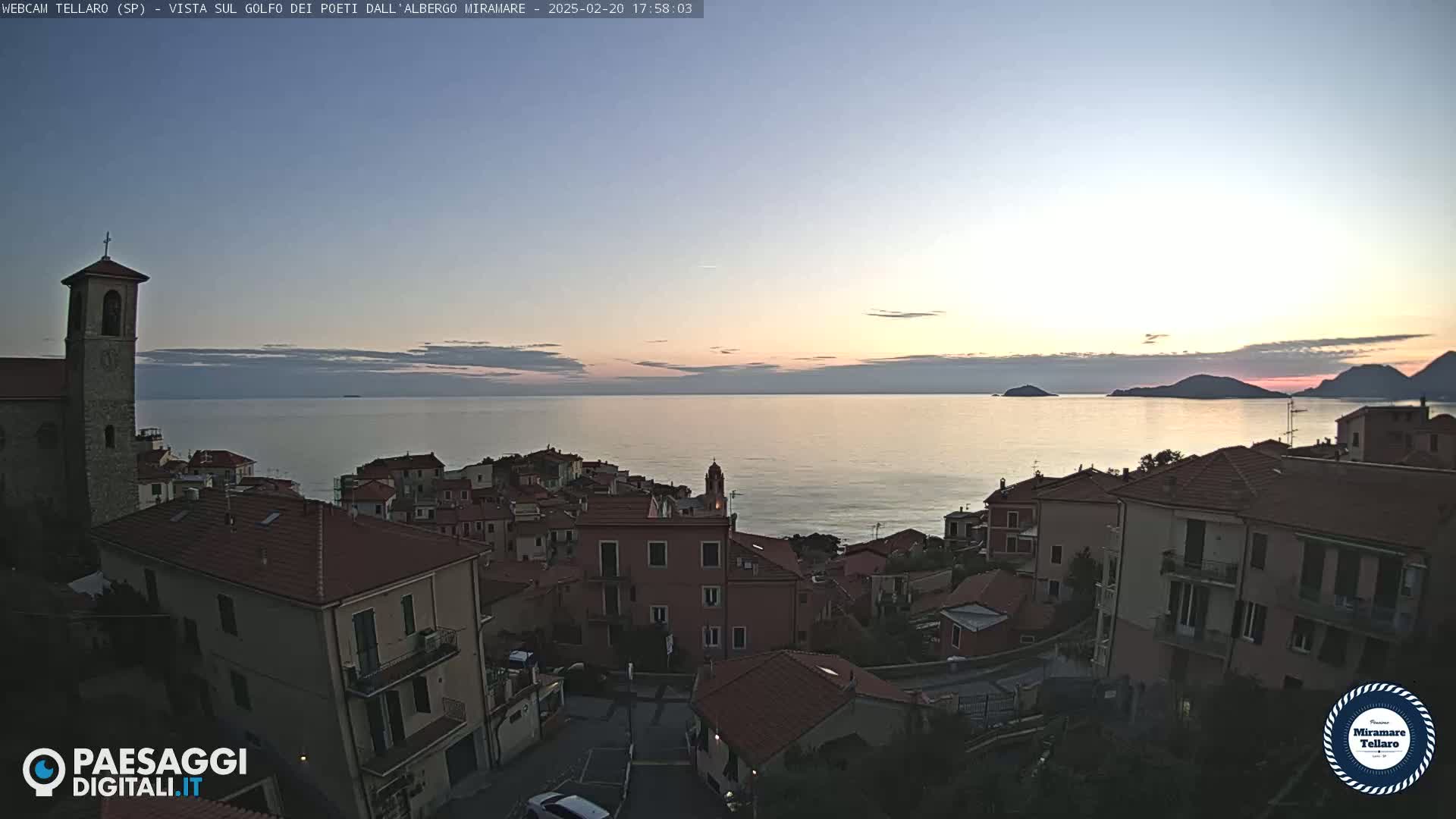A coastal town with red-tiled roofs overlooks a calm sea at sunset under a clear sky.
