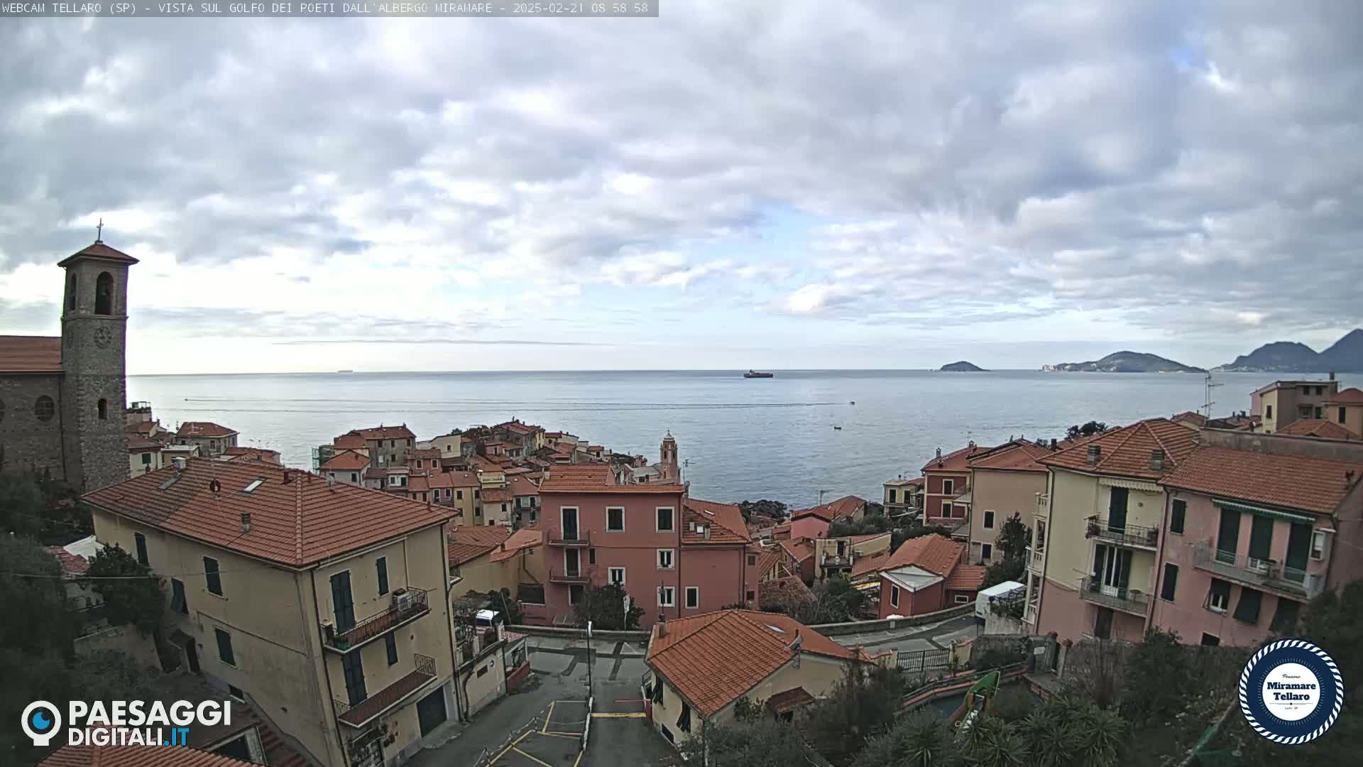 A coastal town with red-tiled buildings overlooks a calm sea under a mostly cloudy sky.