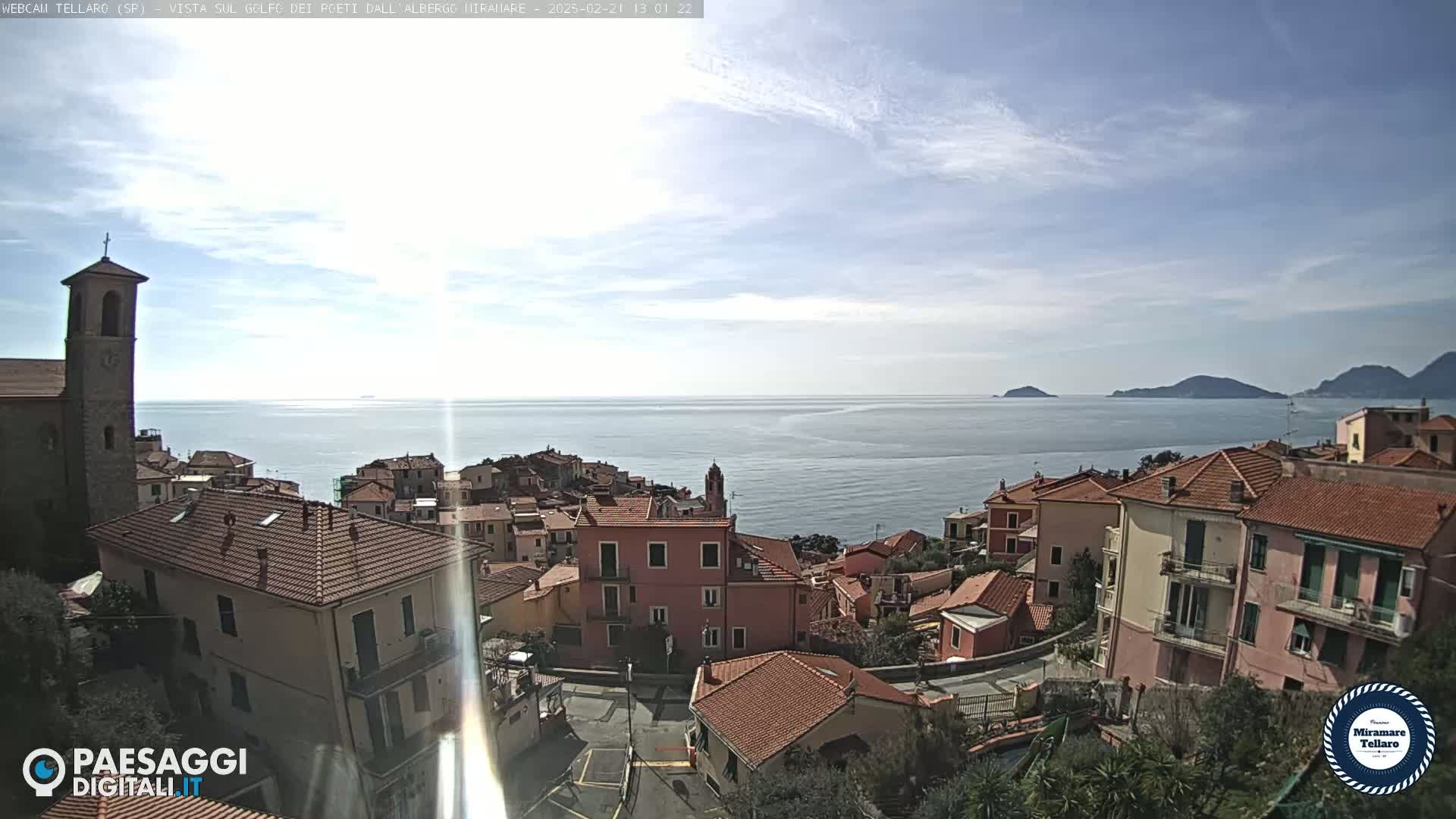A coastal town with red-tiled roofs, a church steeple, and a calm ocean under a mostly sunny sky with scattered clouds.