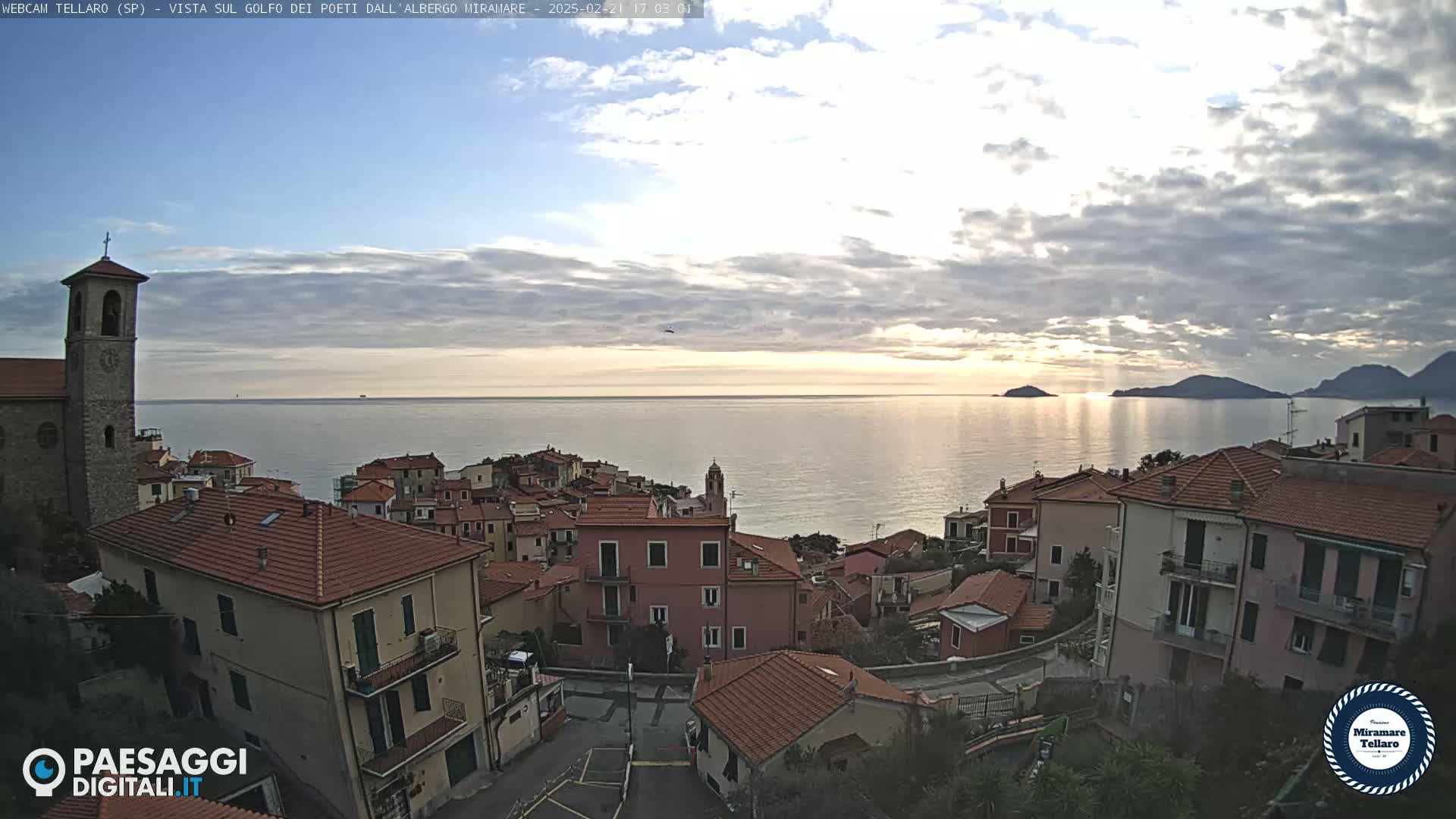 A coastal town with red-tiled rooftops overlooks a calm sea under a partly cloudy sky at sunset.