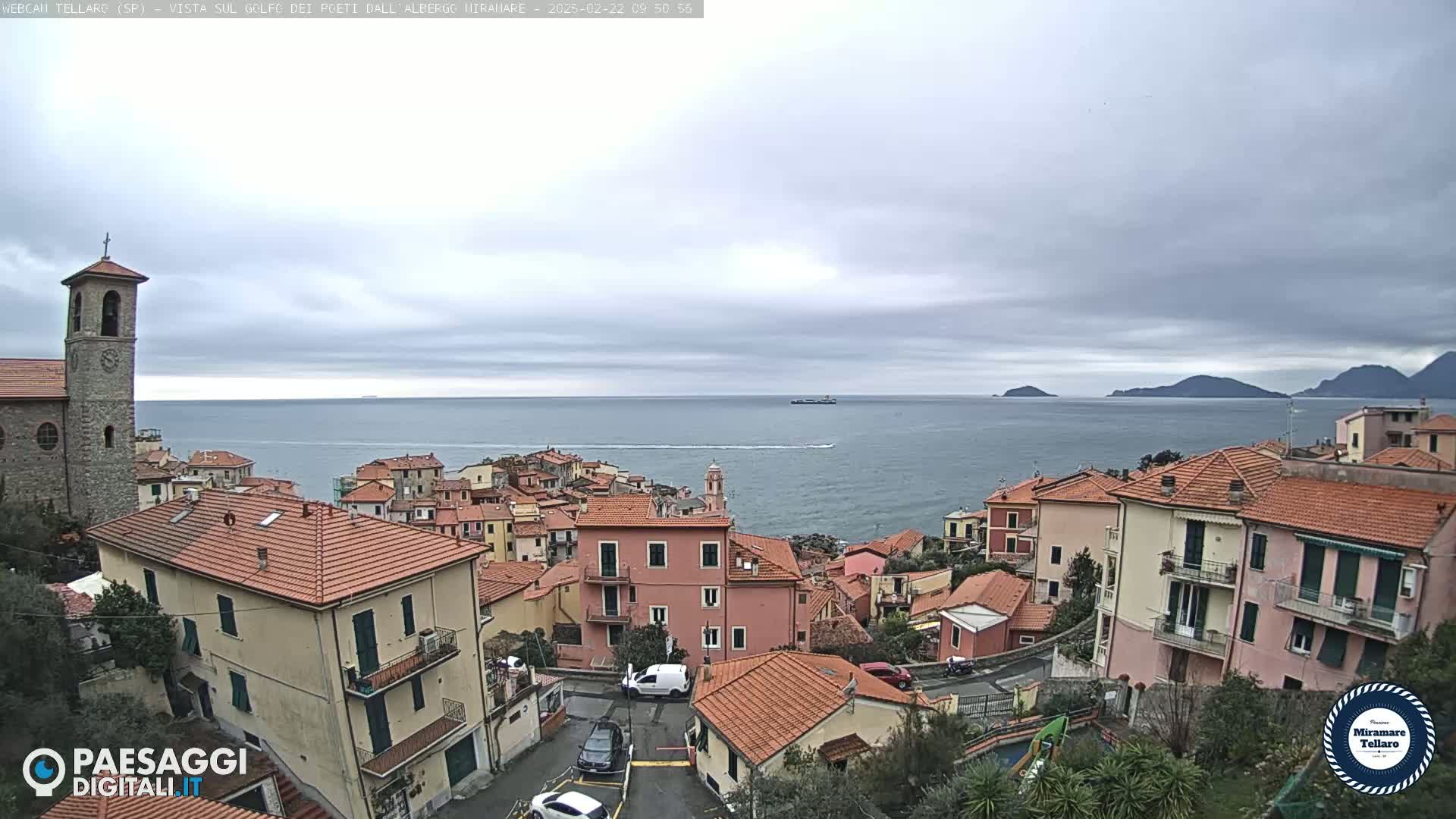 A coastal town with red-tiled roofs and a church overlooks a calm sea under a cloudy sky.