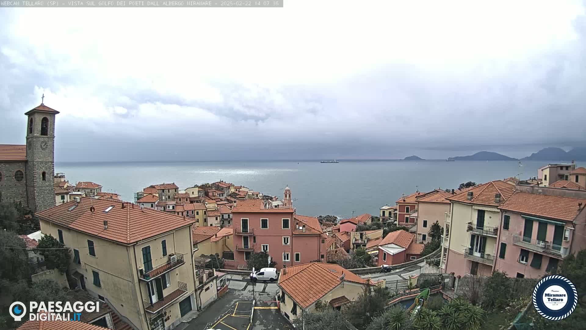 A coastal town with red-tiled roofs and a church steeple overlooks a calm sea under a cloudy sky.