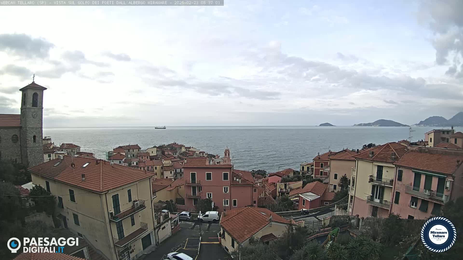 An overcast day shows a coastal town with red-tiled roofs, a church steeple, and a calm sea with distant hills and a single ship.