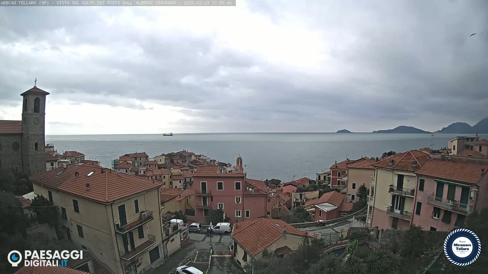 A coastal town with red-tiled roofs and a church overlooks a calm sea under a cloudy sky.