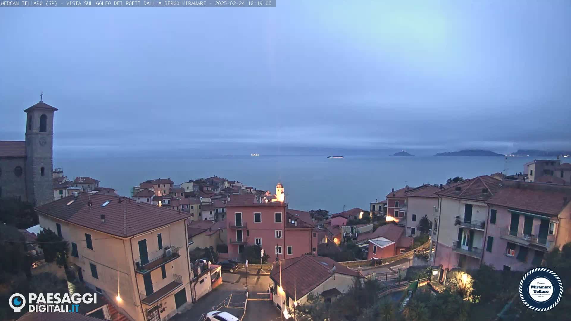 A coastal town with red-tiled roofs overlooks a calm, twilight sea under a cloudy sky.