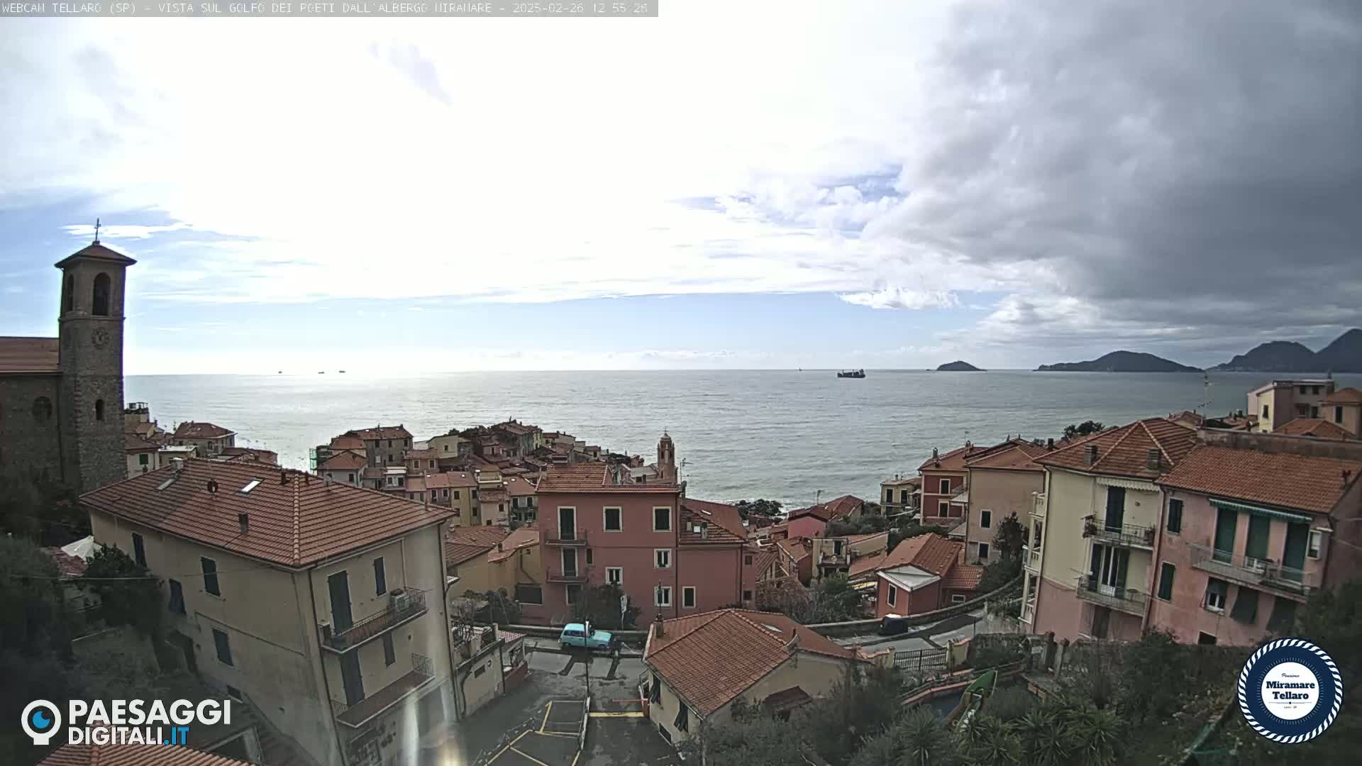 A coastal town with red-tiled roofs and a church steeple overlooks a calm sea under a partly cloudy sky.