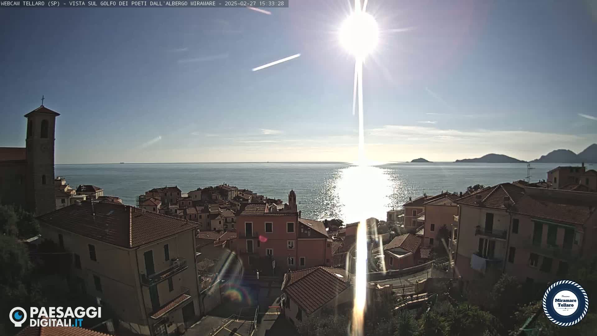 A coastal town with red-tiled roofs overlooks a calm, sun-dappled sea under a bright, sunny sky.