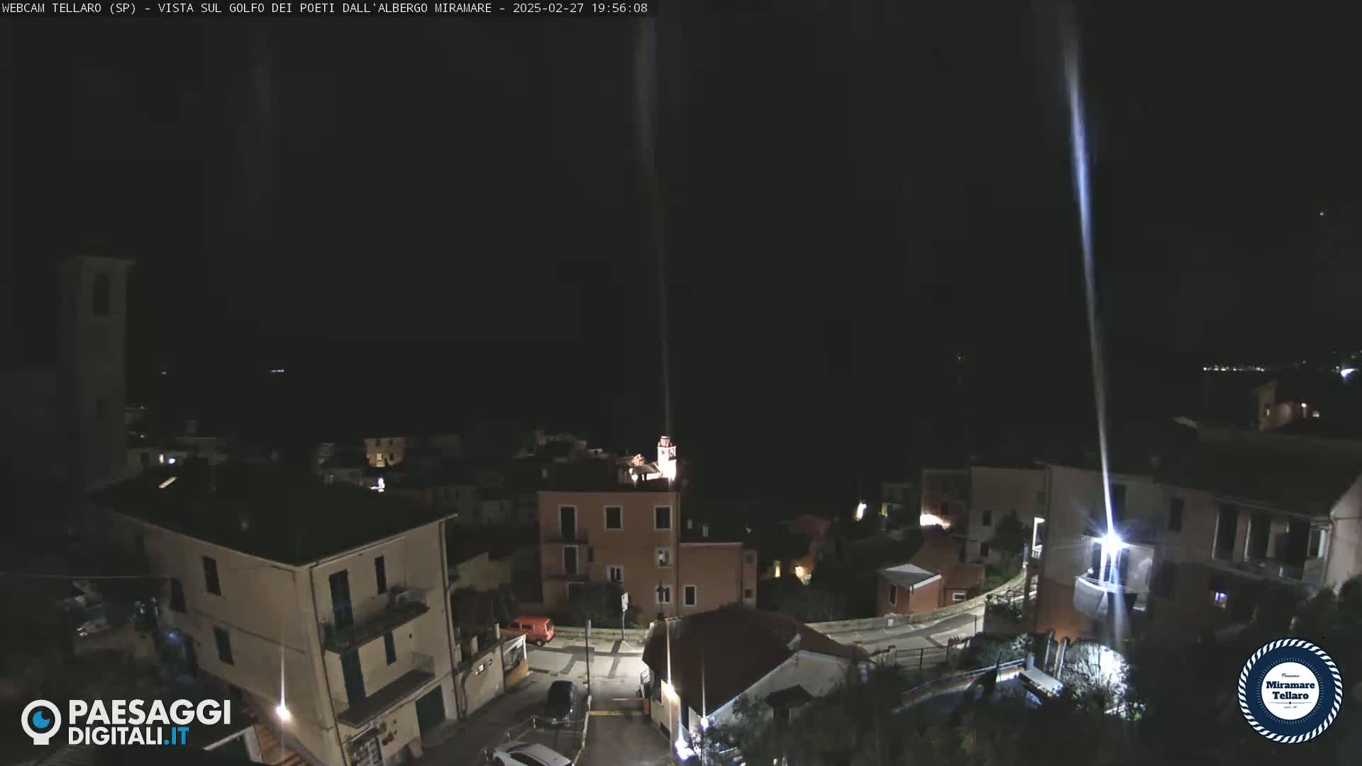 A nighttime, high-angle view of a small town nestled near a dark body of water, illuminated by artificial lights and a single, bright beam of light extending from the ground into the sky.