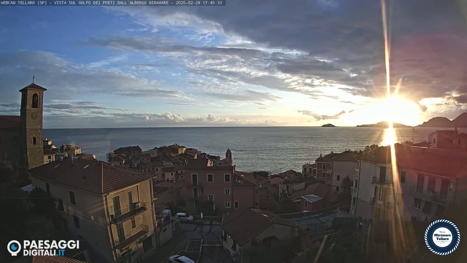A coastal town with terracotta rooftops is nestled against a calm sea under a partly cloudy sunset.