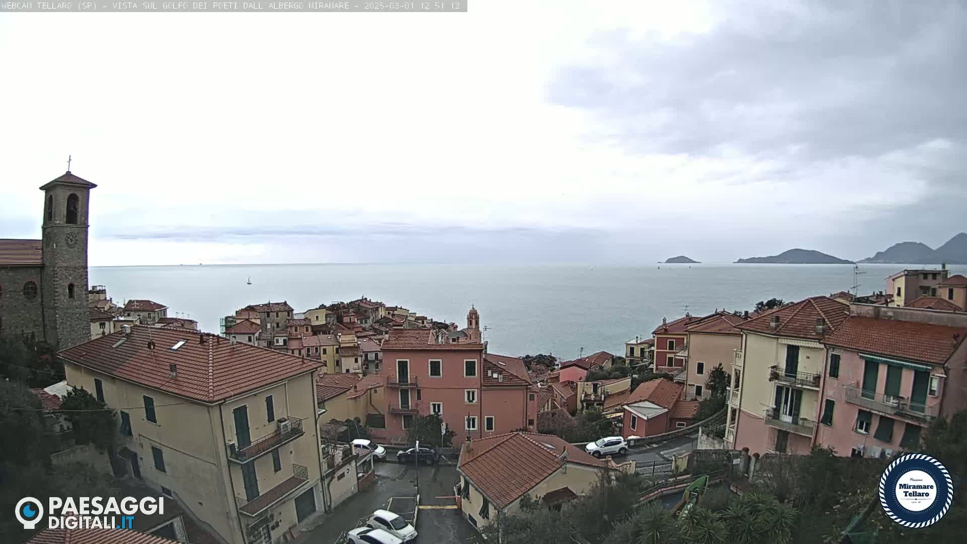 A coastal town with red-tiled roofs and a church steeple overlooks a calm sea under an overcast sky.