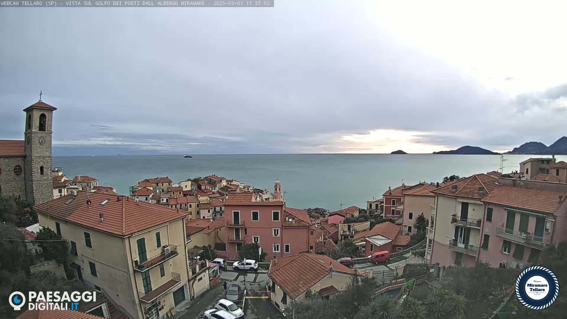A coastal town with red-tiled roofs overlooks a calm sea under an overcast sky.