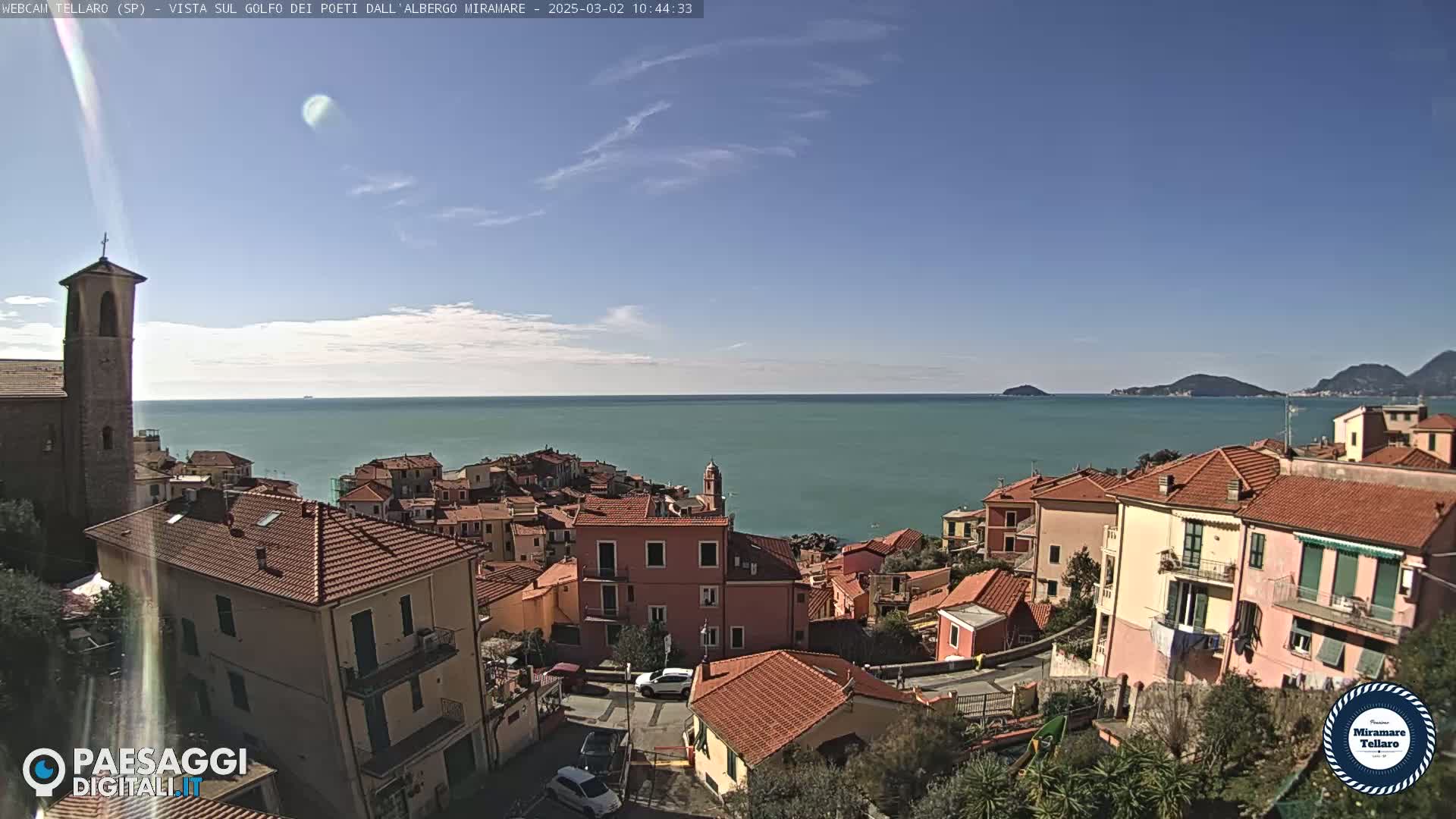 A coastal town with red-tiled roofs overlooks a calm, teal sea under a mostly clear blue sky.