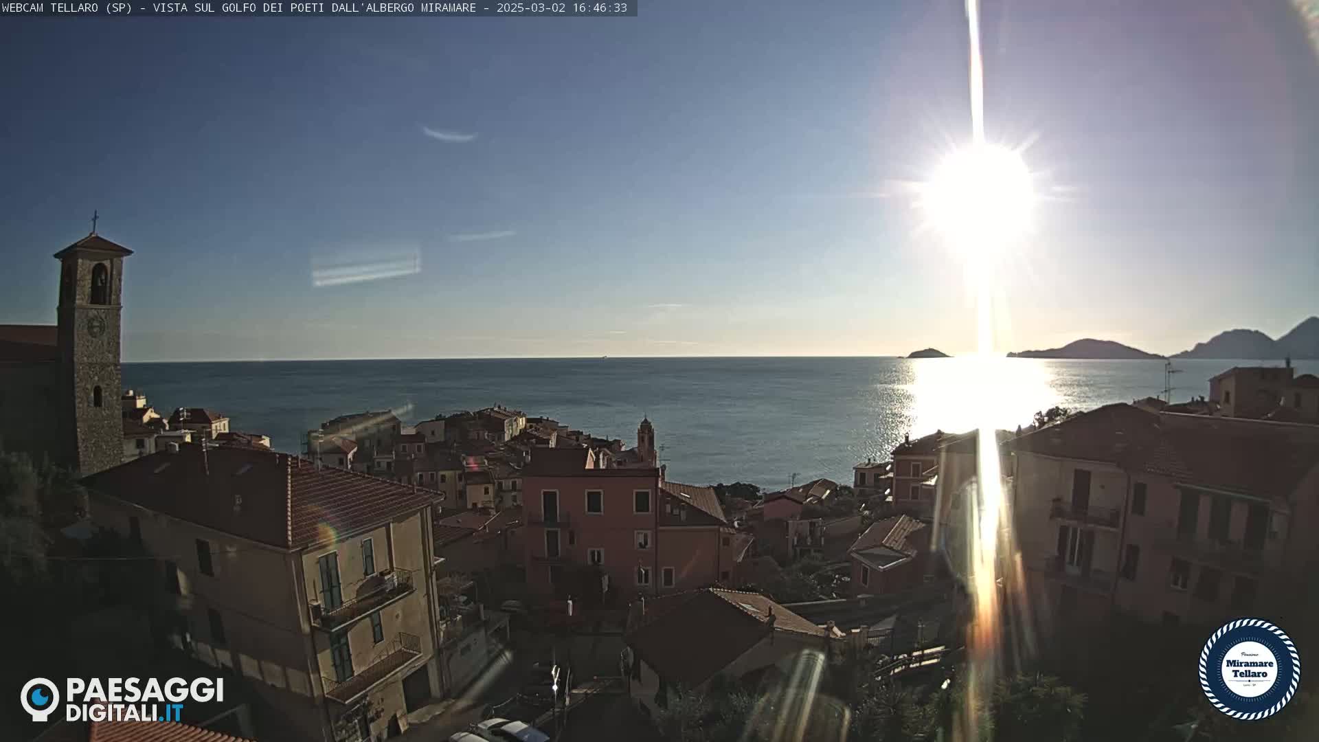 A coastal town with red-roofed buildings is brightly lit by the sun over a calm sea on a clear day.