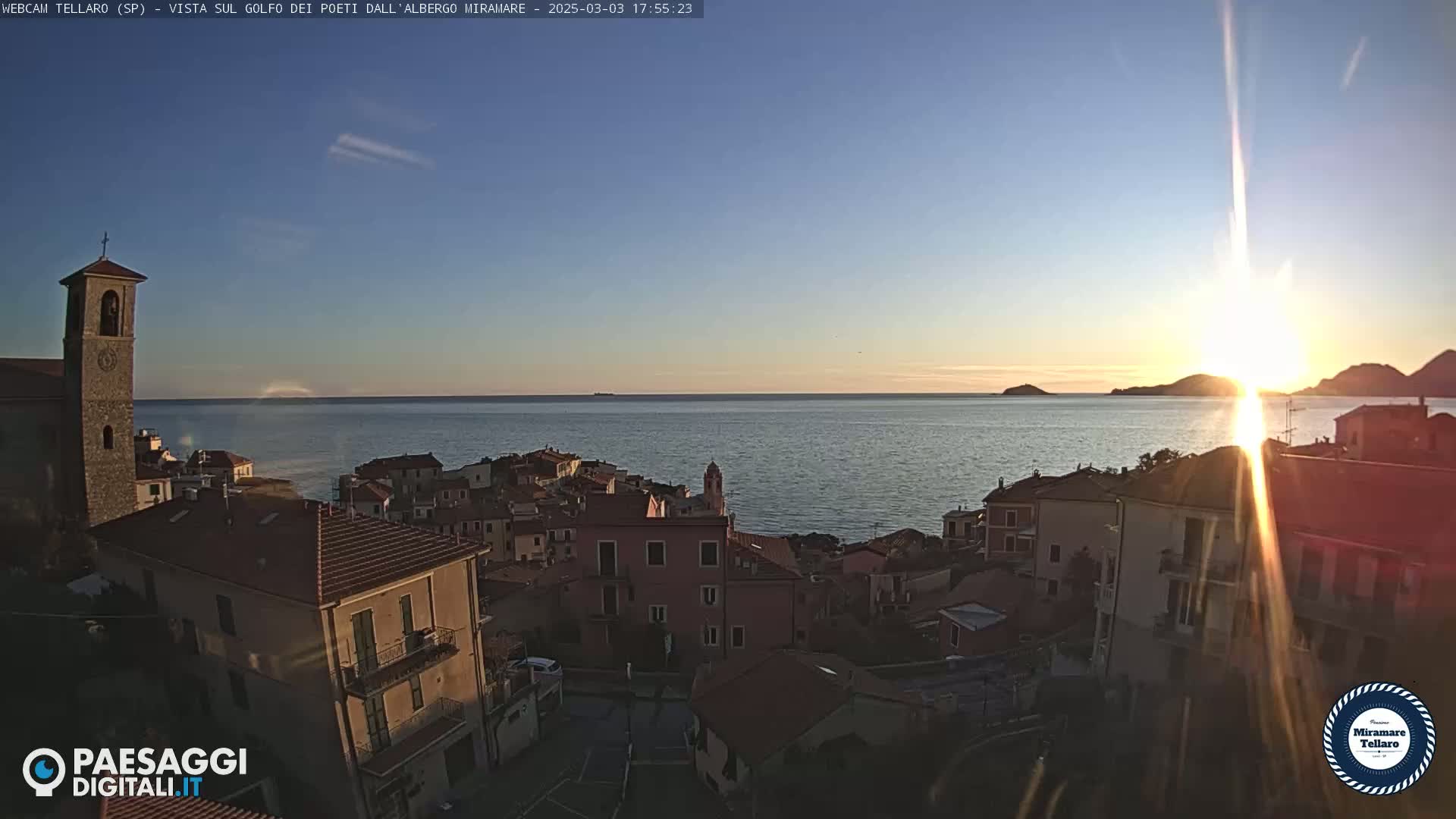 A coastal town with red-tiled roofs and a church steeple is silhouetted against a setting sun over calm water.
