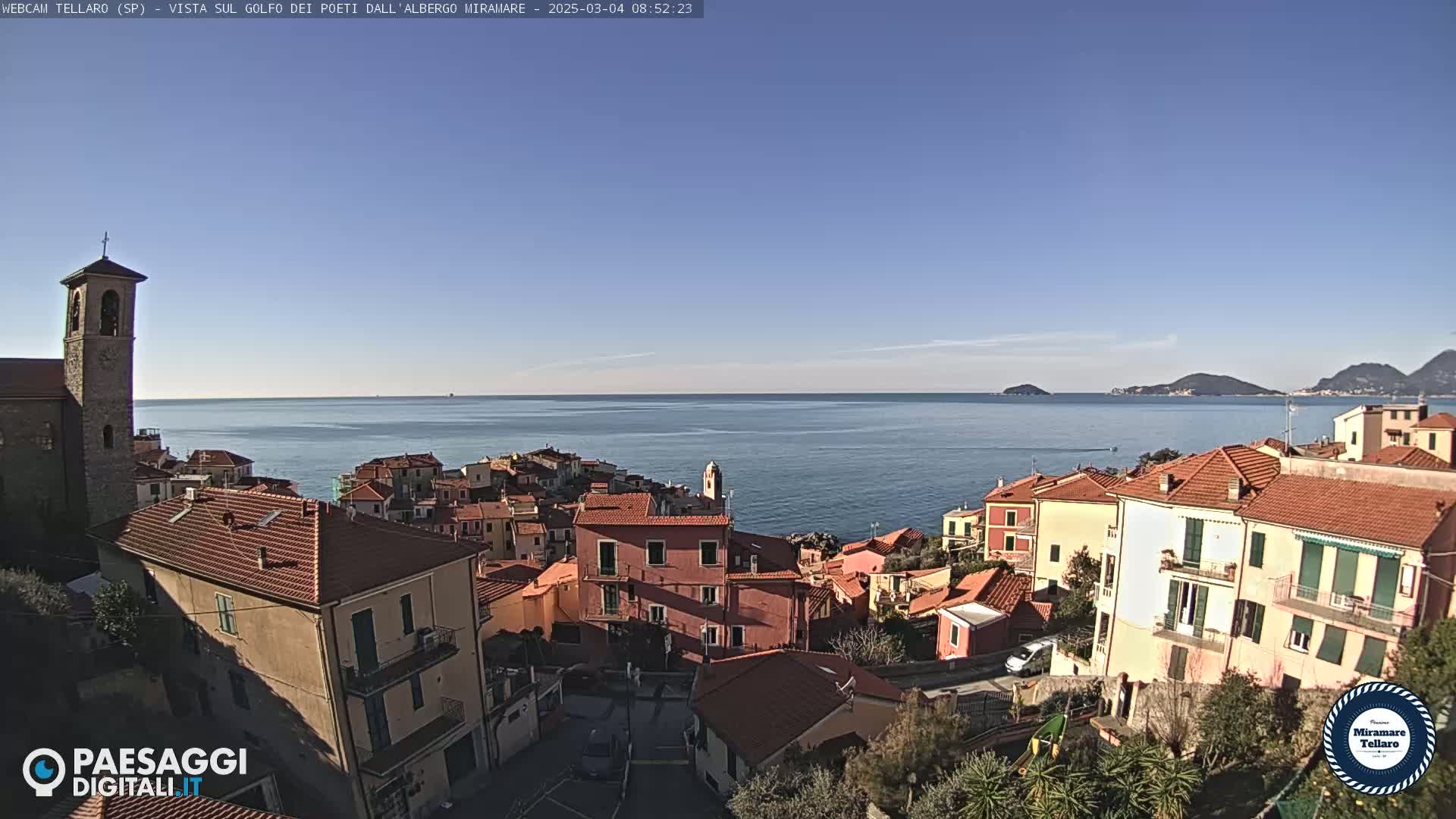 A coastal town with red-roofed buildings overlooks a calm blue sea under a clear, sunny sky.