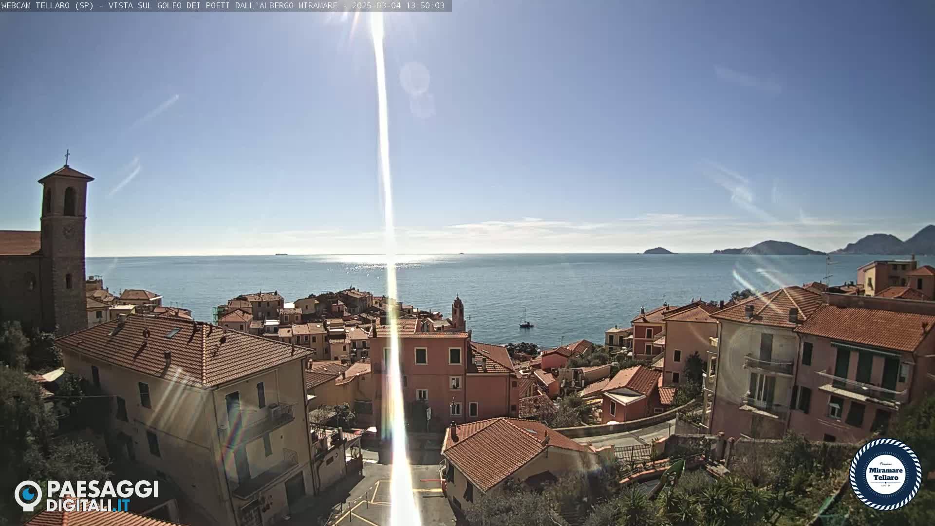 A coastal town with red-tiled roofs overlooks a calm, sunny sea under a clear blue sky.