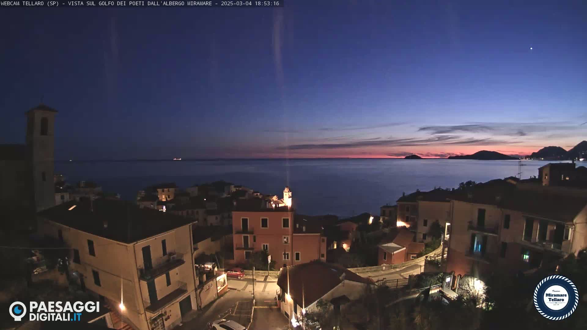 A coastal town at twilight, with the calm sea reflecting the pinkish sunset hues in the sky.