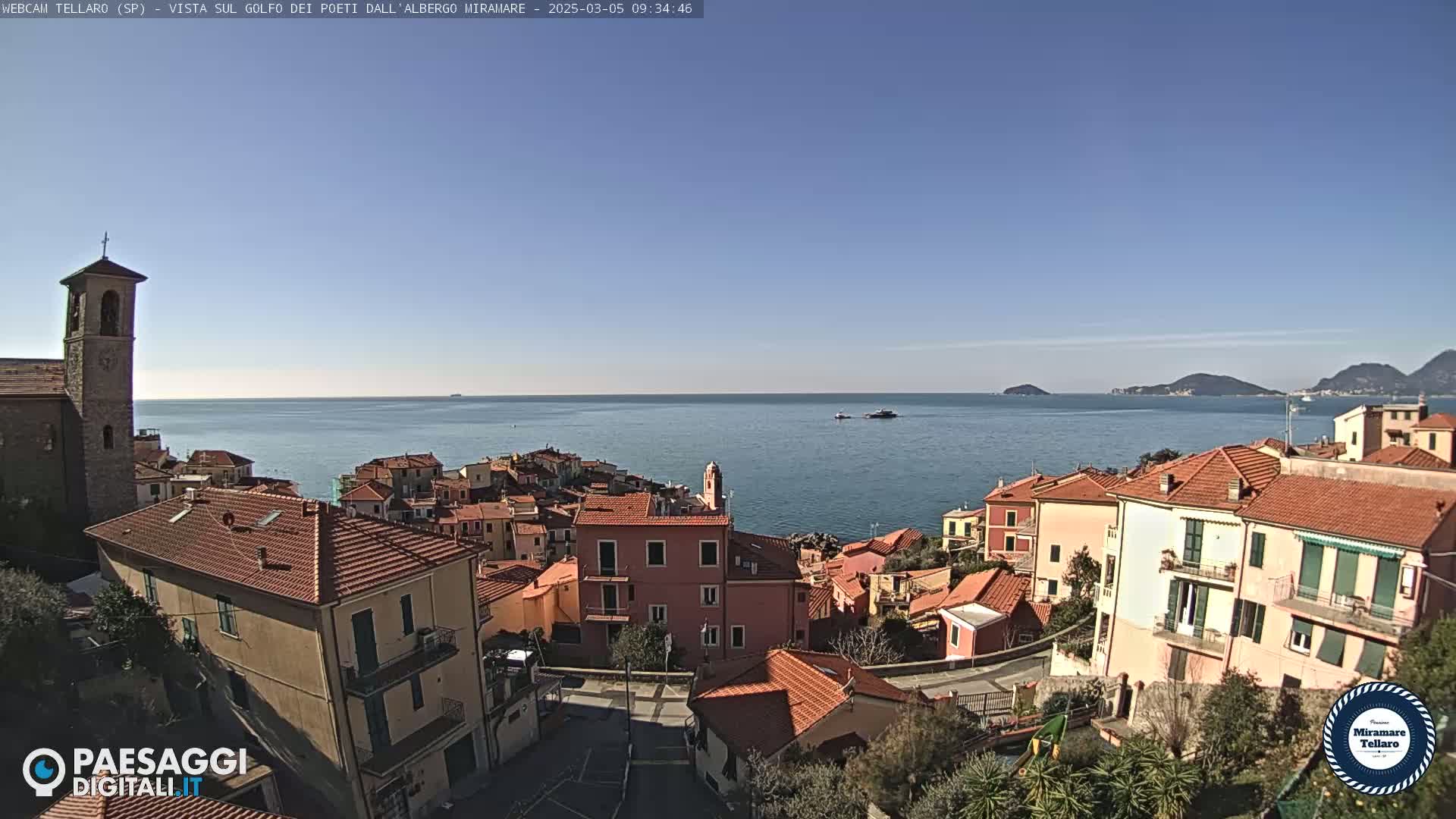 A coastal town with red-tiled buildings, a church steeple, and a clear blue sky overlooks a calm sea with distant islands.