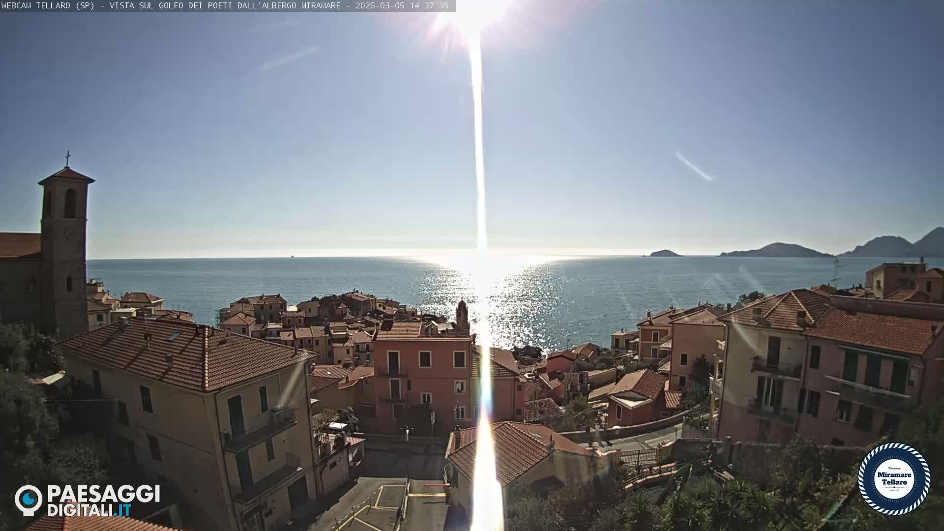 A sunlit coastal town with red-tiled roofs overlooks a calm sea under a clear blue sky.