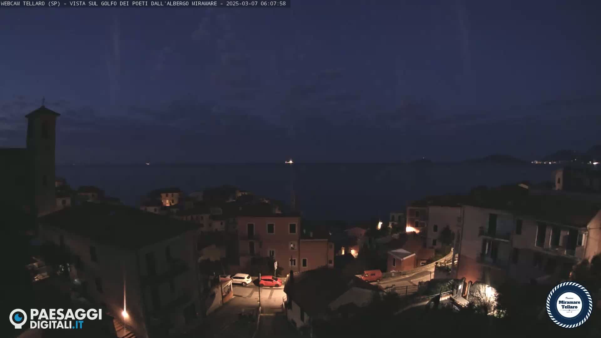 A dark, nighttime view of a coastal town overlooking a calm sea under a mostly clear sky.