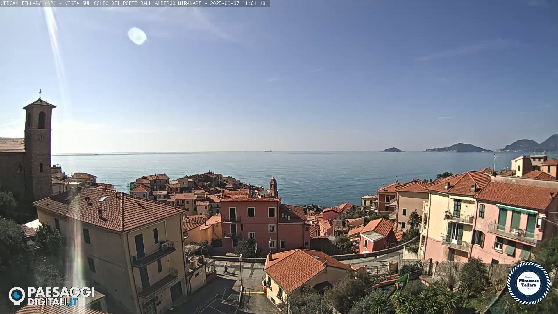 A coastal town with red-tiled roofs and a church overlooks a calm blue sea under a clear sky.