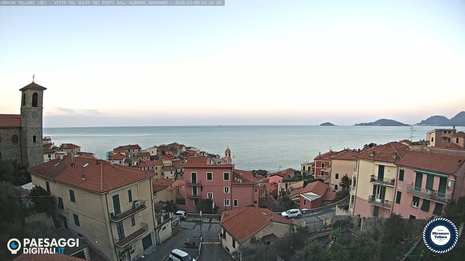 A coastal town with red-tiled roofs and a church steeple overlooks a calm sea under a clear, light-colored sky.