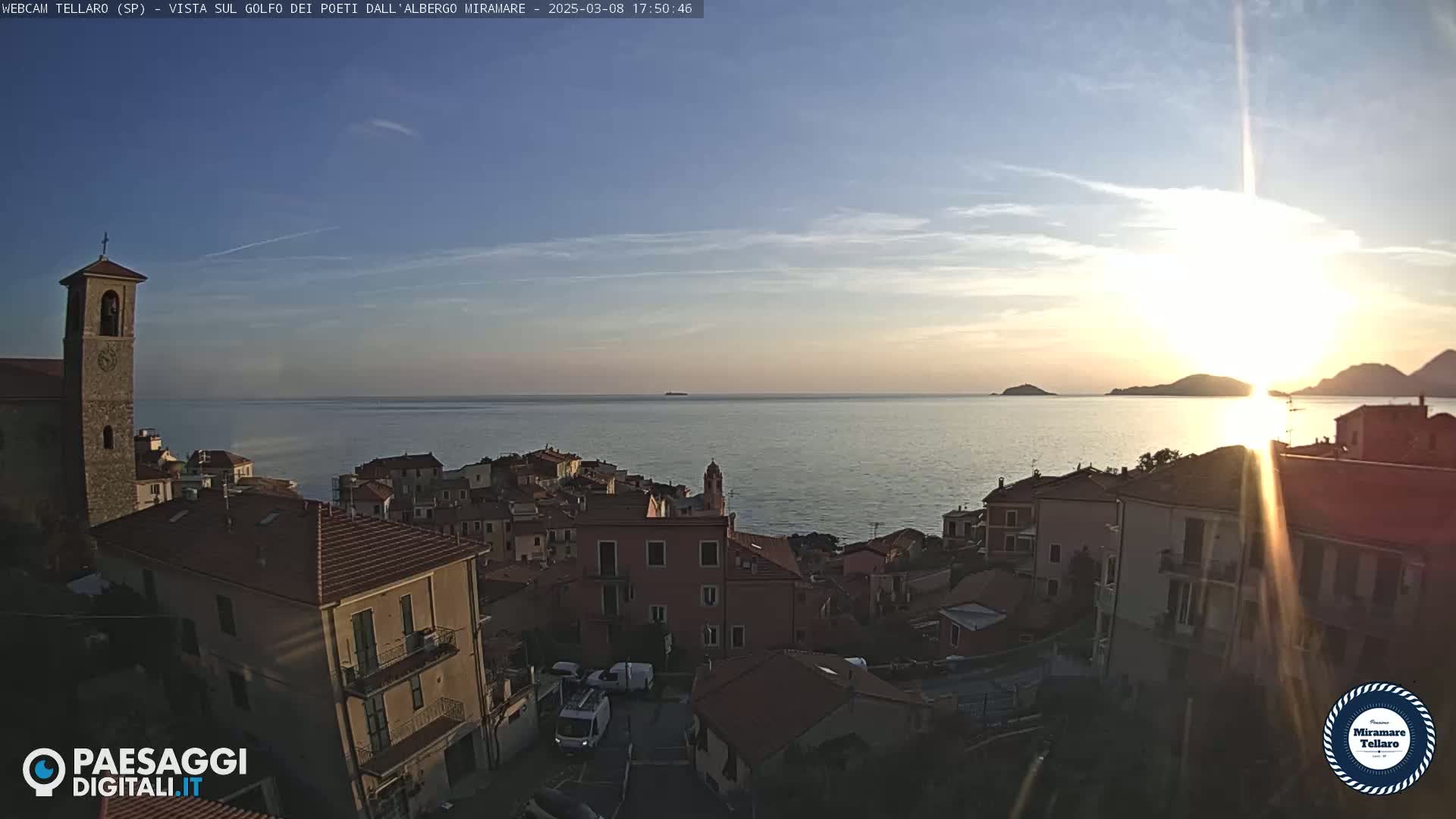 A coastal town with red-tiled roofs sits on a hillside overlooking a calm sea at sunset.