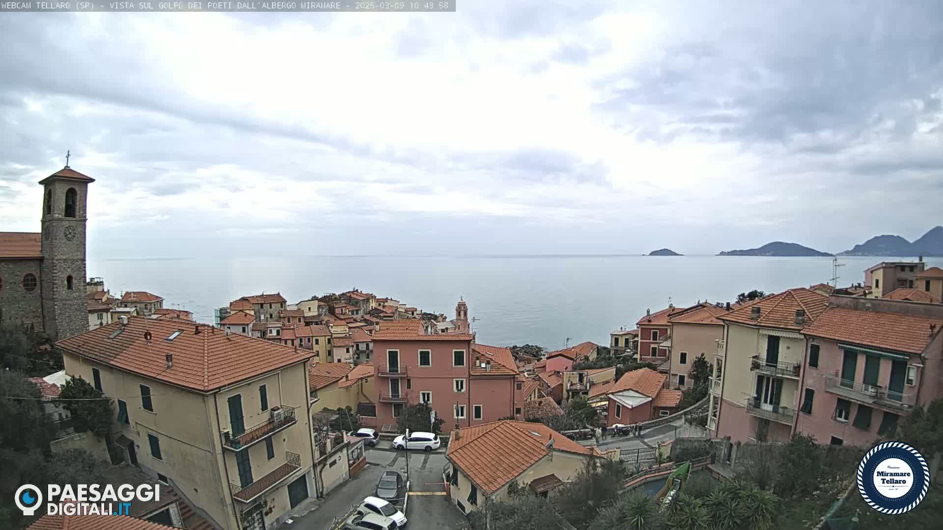 A coastal town with red-tiled roofs overlooks a calm sea under a mostly cloudy sky.