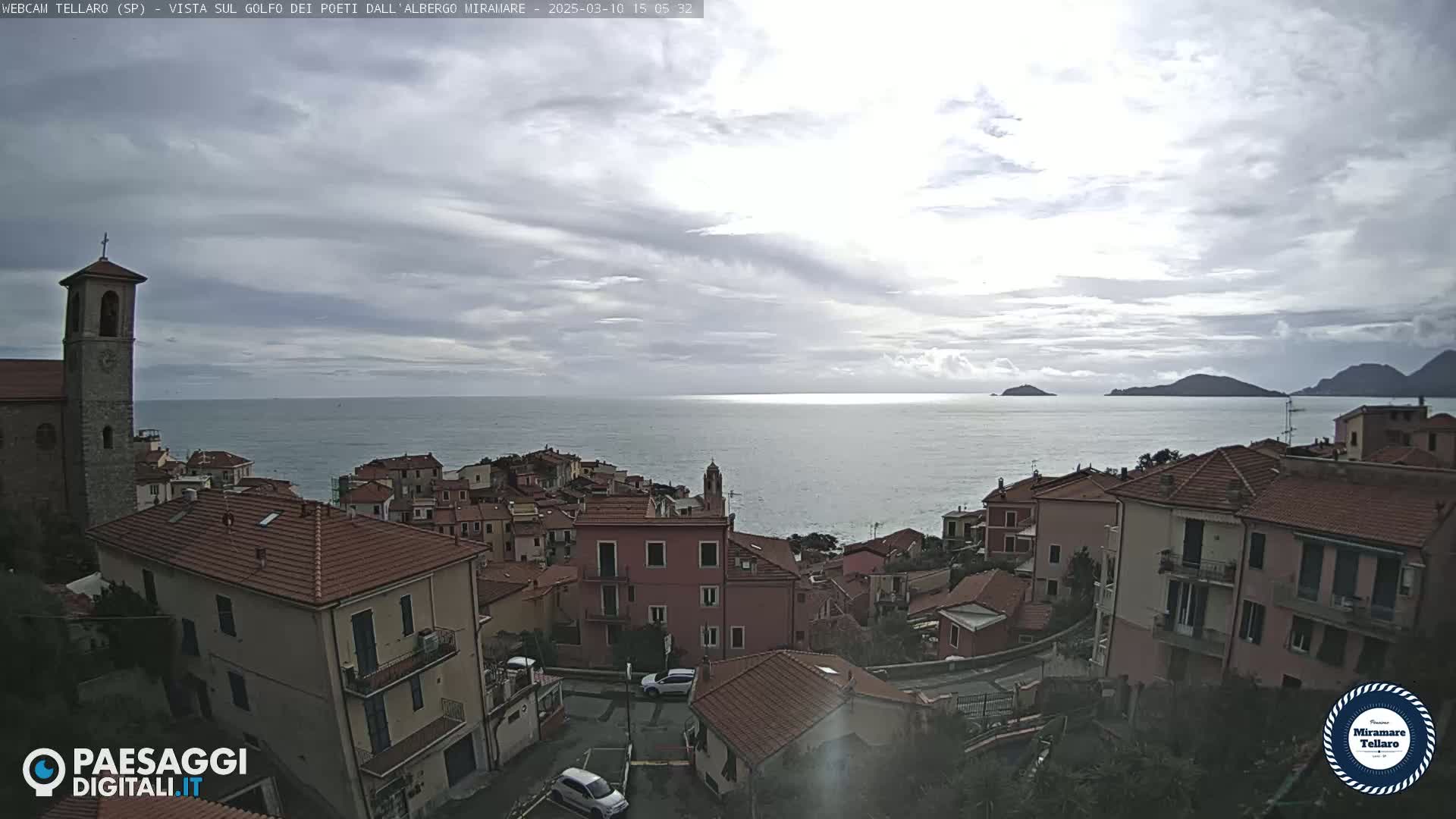 A coastal town with red-tiled roofs overlooks a calm sea under a mostly cloudy sky.