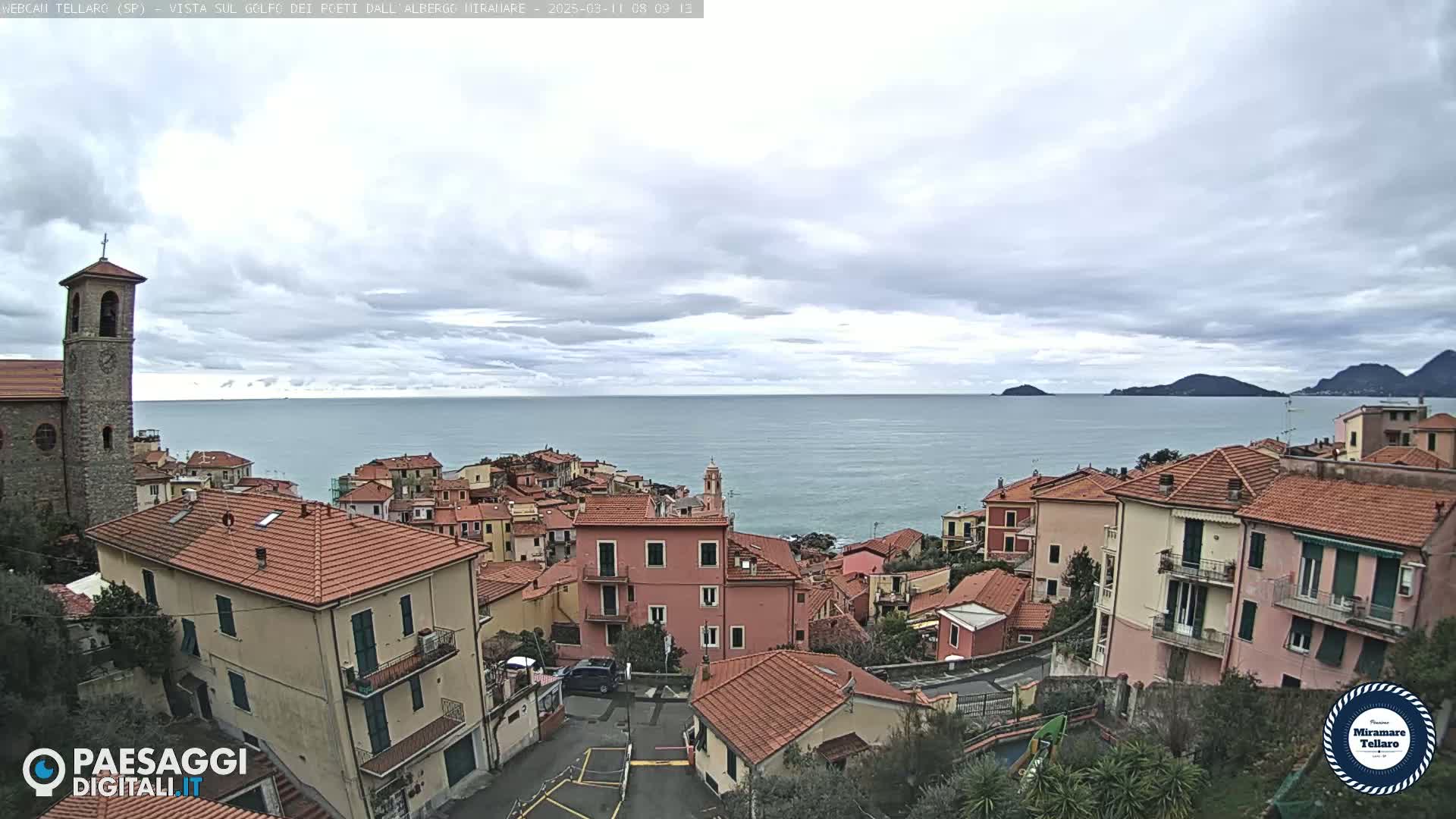 A coastal town with red-tiled roofs overlooks a calm sea under a cloudy sky.