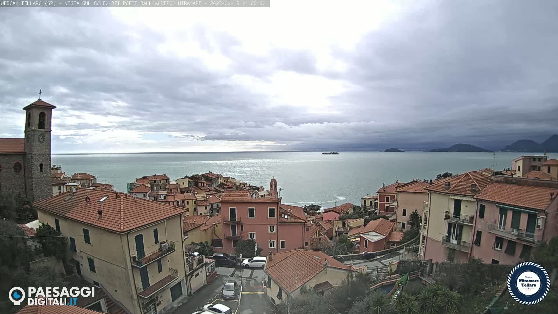 A coastal town with red-tiled roofs and a church steeple overlooks a calm sea under a cloudy sky.