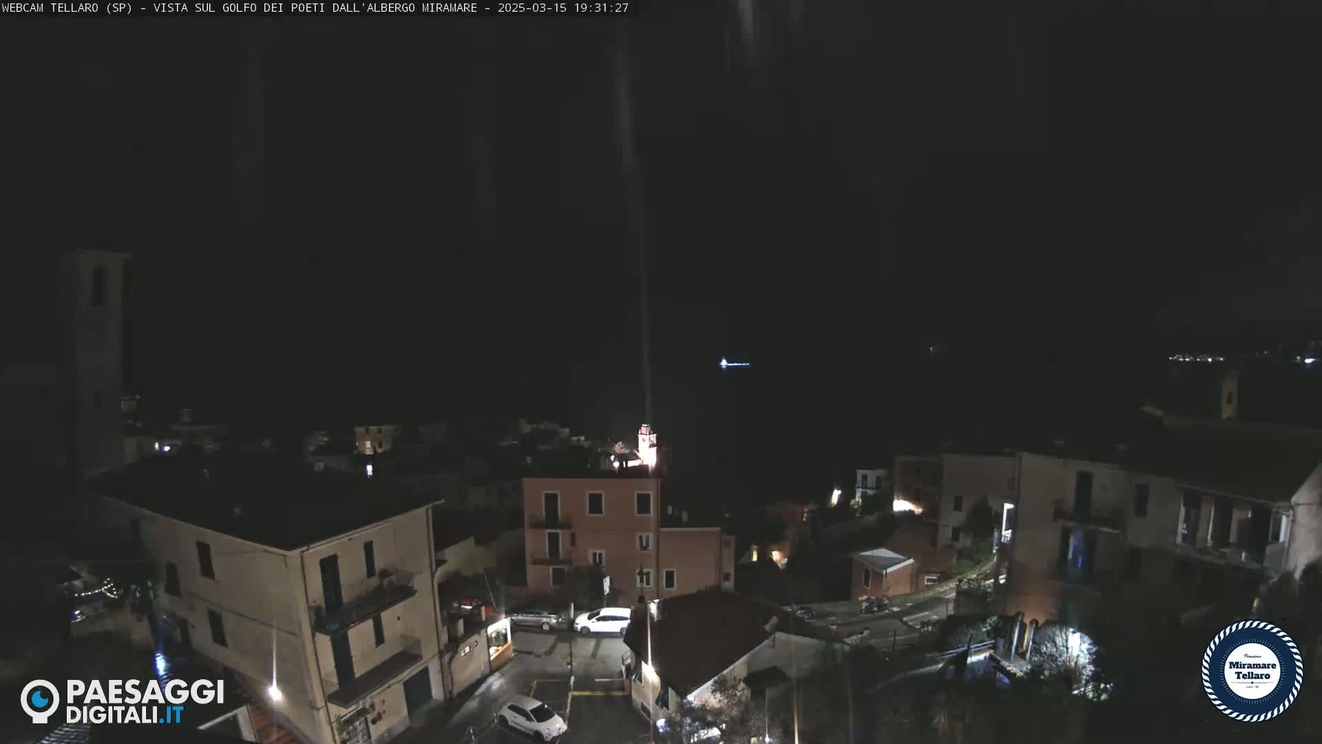 A nighttime, high-angle view of a small town nestled on a hillside, with a dark sky and faintly visible lights in the distance.
