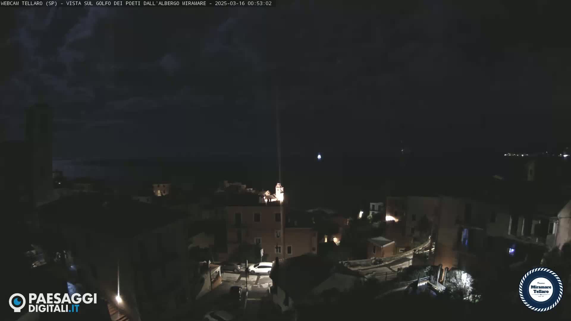 A nighttime, overhead view of a coastal town with dimly lit buildings, a dark ocean in the background, and a mostly clear sky.