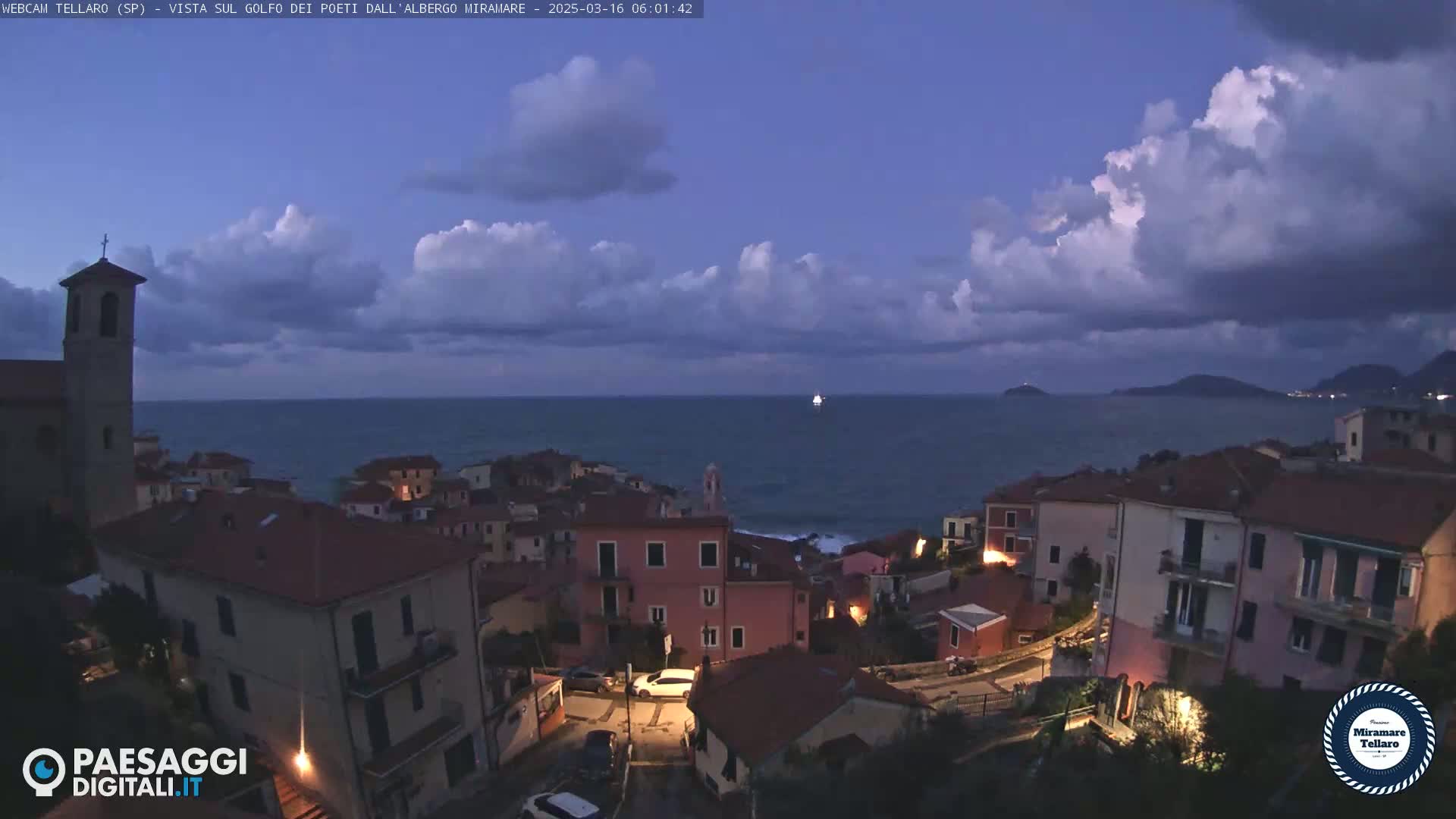 A coastal town with a church is illuminated at twilight under a partly cloudy sky, overlooking a dark sea with a single ship visible in the distance.