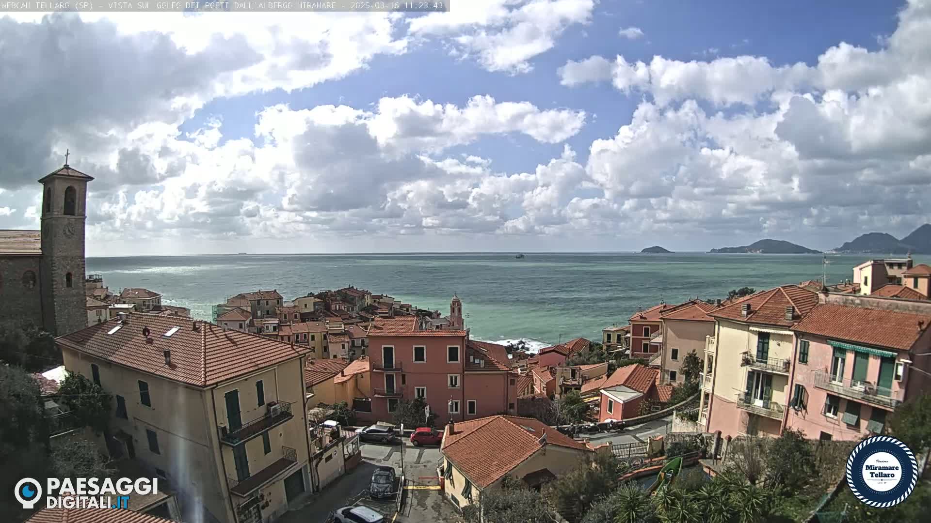 A coastal town with red-tiled buildings overlooks a calm sea under a partly cloudy sky.